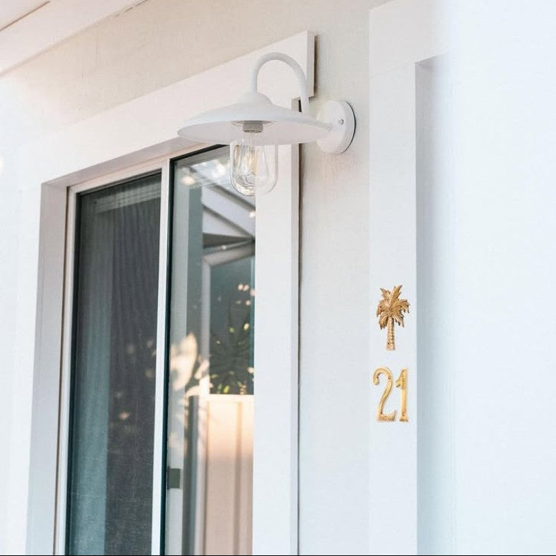 A white outdoor wall sconce light fixture mounted on a white exterior wall next to a glass sliding door, with a brass gold house number 21 and braa palm tree plaque on the pillar.