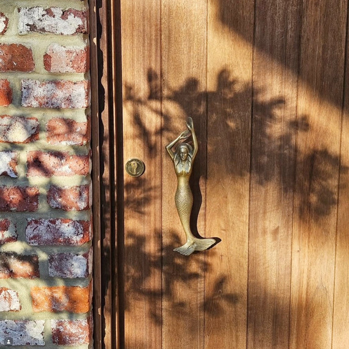 A bronze mermaid door handle shaped like a mermaid and raised arms, mounted on a wooden door next to a brick wall, with dappled sunlight and leaf shadows cast across the surface.