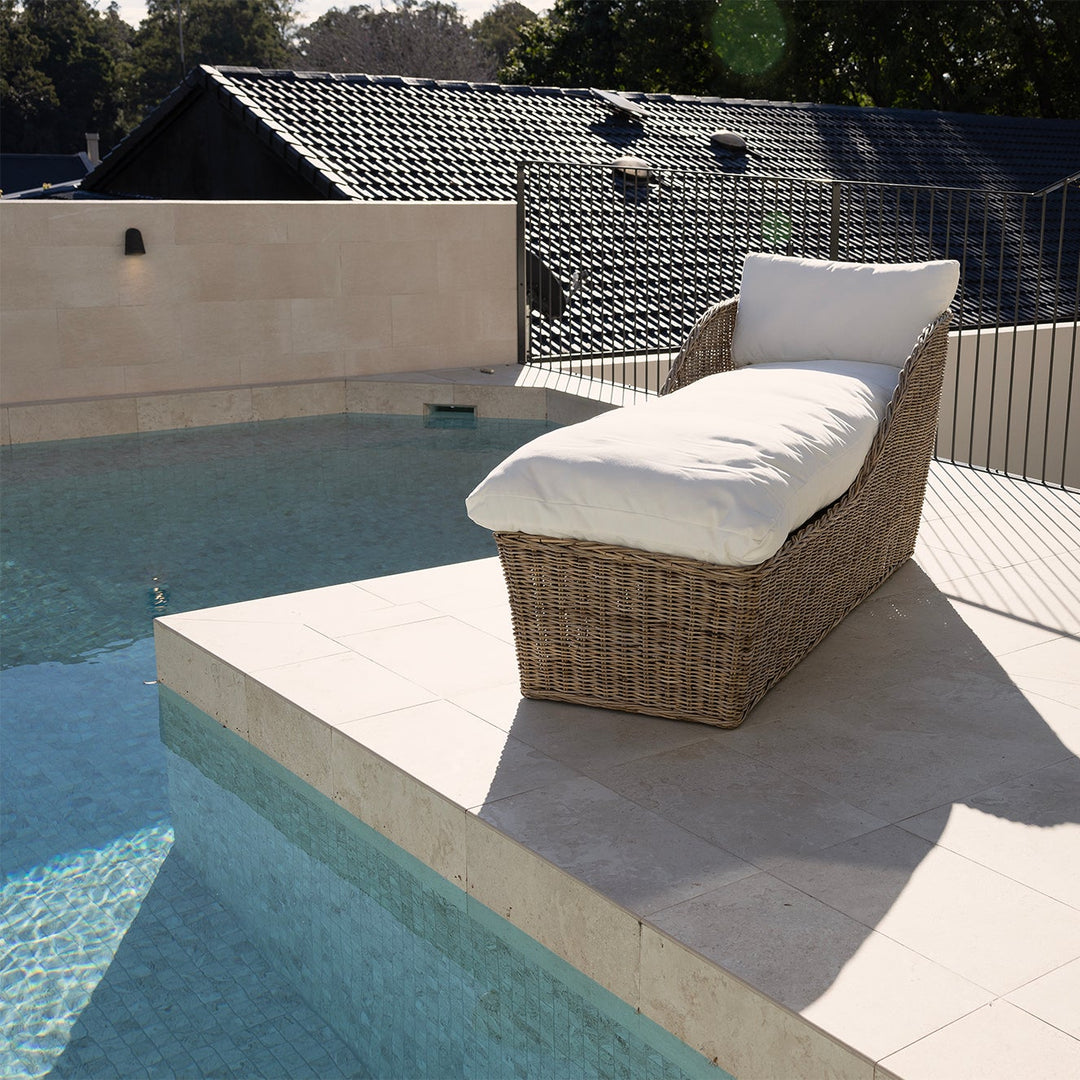 A rattan outdoor chaise lounge chair with white cushions sits on a tiled poolside patio next to a swimming pool.
