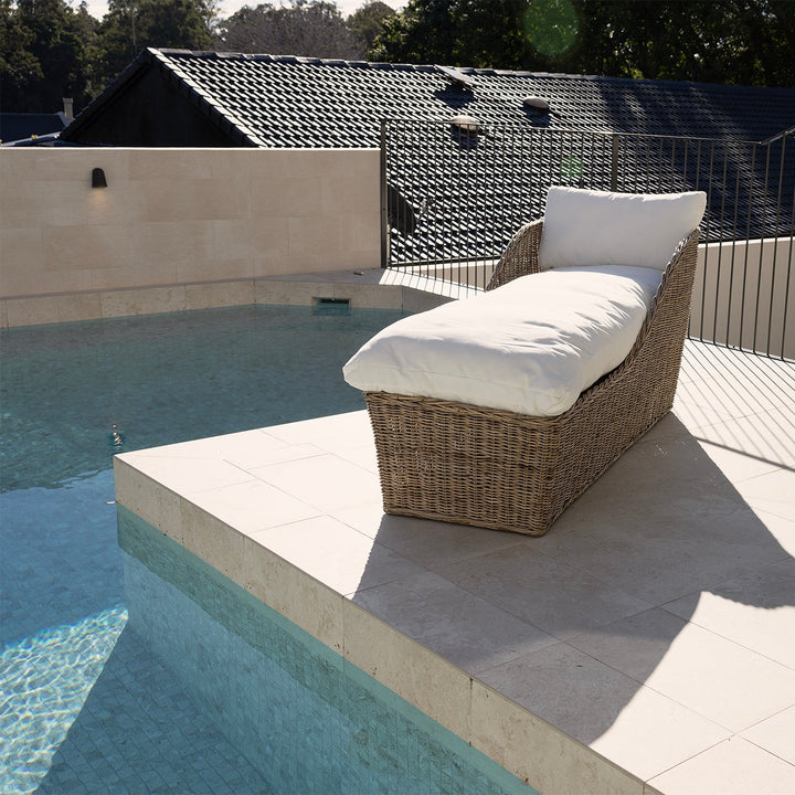 A rattan outdoor chaise lounge chair with white cushions sits on a tiled poolside patio next to a swimming pool.