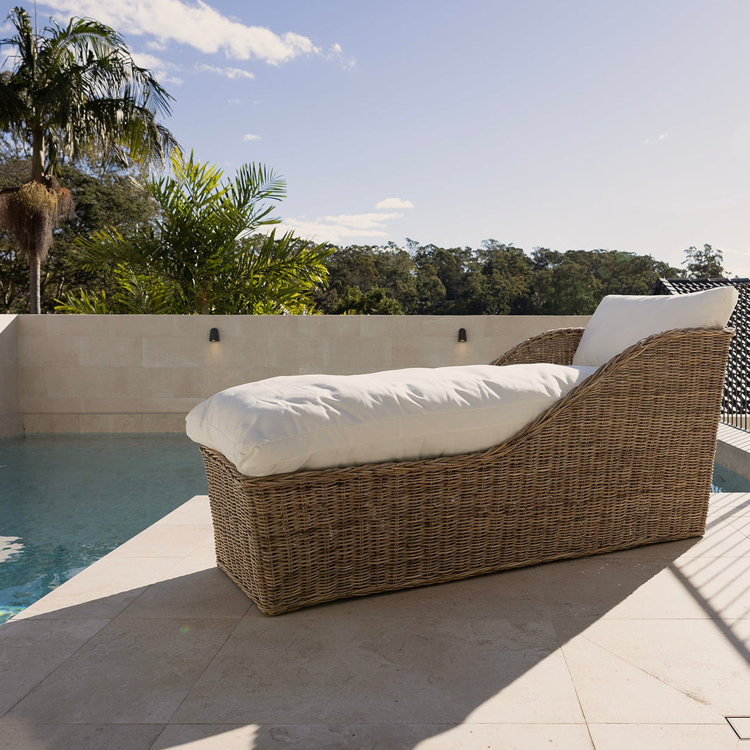 A rattan outdoor chaise lounge chair with white cushions sits beside a swimming pool on a tiled patio, with a metal fence, trees, and sky in the background.