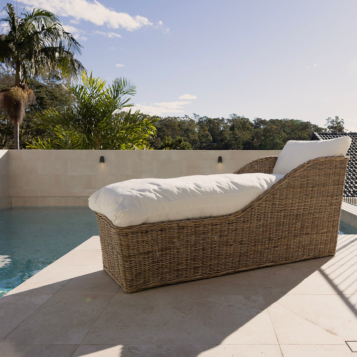 A rattan outdoor chaise lounge chair with white cushions sits beside a swimming pool on a tiled patio, with a metal fence, trees, and sky in the background.