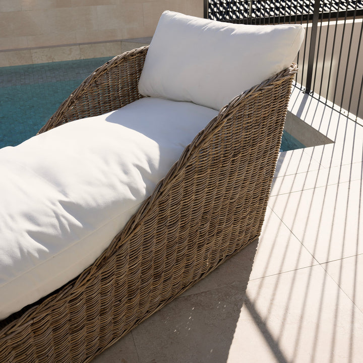A rattan chaise lounge chair with thick white cushions, positioned on a tiled outdoor patio next to a swimming pool.