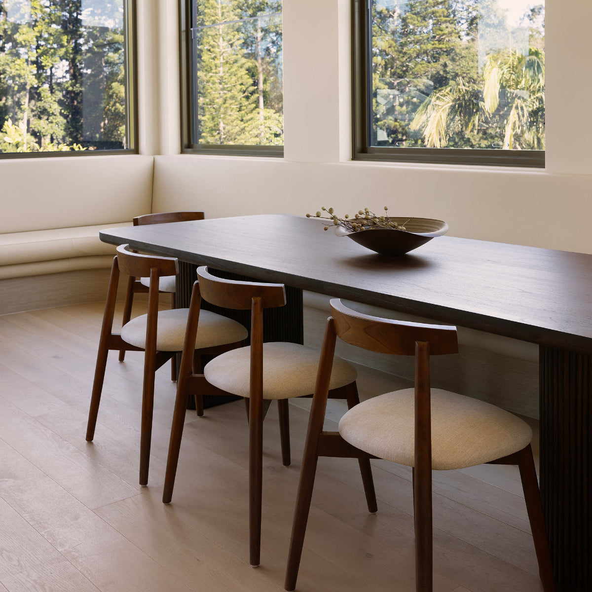 Dining area with a wooden table and chairs in front of large windows with a view of trees by Ocean Luxe