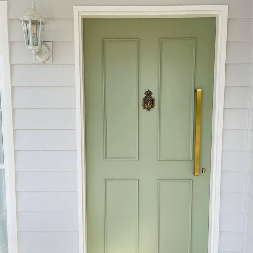 A sage green paneled front door with a decorative brass pineapple door knocker, set within a white frame on a white shiplap wall with a white wall sconce.